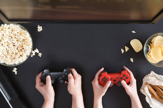 Man and woman playing video games together, top view of gamer accessorises and snacks frame, flat lay on black background. Joystick and gamepad, keyboard, game console, beer, chips and popcorn.