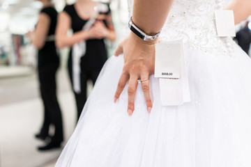 Closeup back of young woman trying on wedding dress in boutique discount store with shiny tulle, expensive dollars price tag, engagement ring, fitness watch