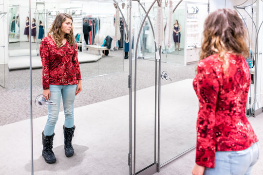 Young woman trying on clothing jeans in boutique store looking at herself in mirror with red shirt and boots