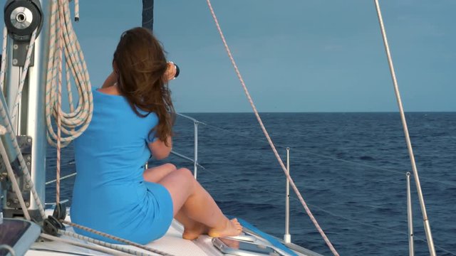 Womanin a blue dress sits aboard the yacht and exploring the horizon through binoculars