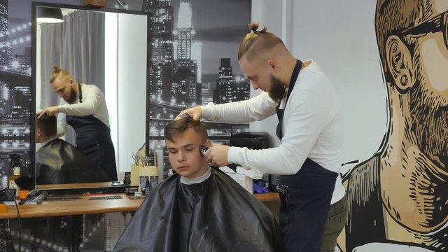 Hairdresser for men. Barbershop. A young guy gets a haircut and hair care service from a bearded man with a hair tied on his head. Hair cutting on the side of the young guy's head with a hair clipper