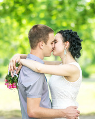 Portrait of beautiful young couple female bride with small wedding pink flowers roses bouquet and male bridegroom kissing in summer park