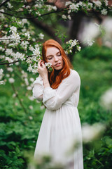 Beautiful red-haired girl in a white dress among blossoming apple-trees in the garden.
