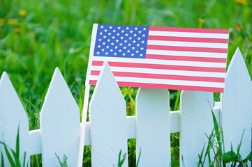 The US flag on a white fence against the background of a blooming green grass.