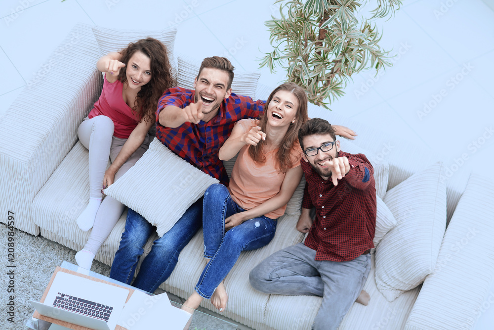 Canvas Prints group of laughing friends sitting on sofa and showing forefinger on the camera.