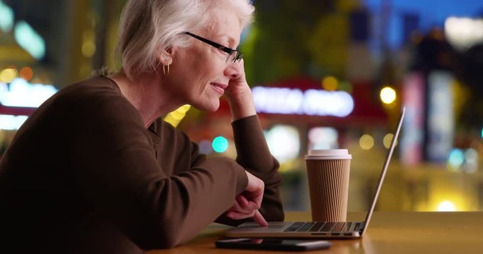 Mature woman sitting at outdoor cafe on city street at night using laptop computer, Senior wearing eyeglasses working on notebook pc on restaurant patio, 4k