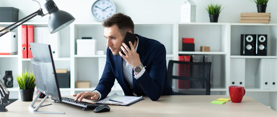 A young man is standing near a table in the office, talking on the phone and typing text on the...