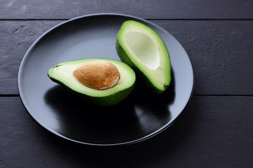 Avocado on a black plate, halves of avocado on a black wooden background, green tropical fruit top...