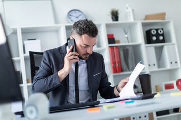 Fototapeta premium A man is sitting in the office at the table, talking on the phone and flipping through documents.
