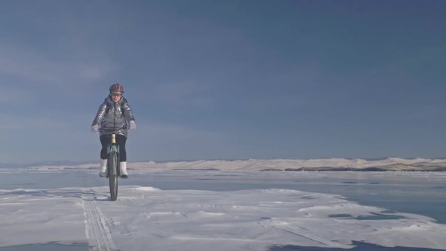 Woman is riding bicycle on the ice. The girl is dressed in a silvery down jacket, cycling backpack and helmet. Ice of the frozen Lake Baikal. The tires on the bicycle are covered with special spikes