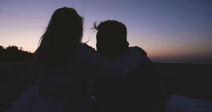 Young couple in love (guy and girl) are sitting on the sand on the sea, sunset, darkness.