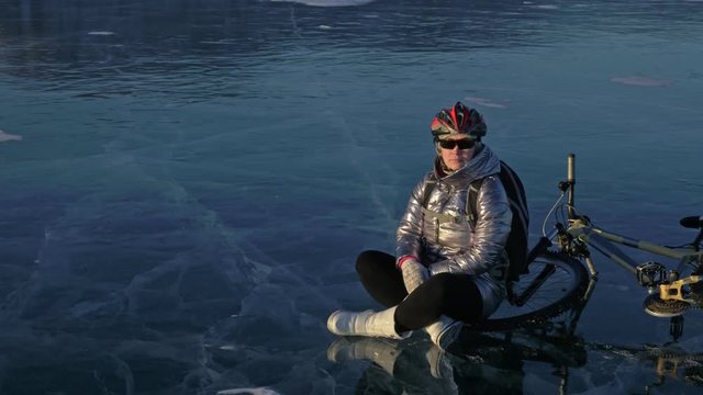 Woman is riding bicycle on the ice. The girl is dressed in a silvery down jacket, cycling backpack and helmet. The cyclist stopped to rest. She sits on the wheel and enjoys the beautiful view of the