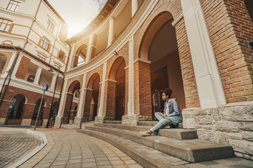 Young curly cute African-American girl with curly Afro hair and in denim overalls is sitting on the stair under a brick arc and waiting for her friend, with copy space place on the left, wide-angle