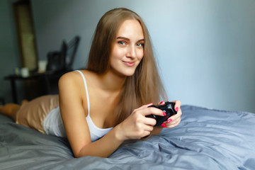 Girl gamer plays with a wireless gamepad while looking at the screen in front of her. Young blonde girl smiles and enjoys winning with gamepad in hand, playing video games console, lying on the bed.