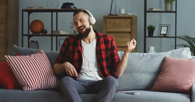 Attractive male hipster in the casual style in the big white headphones listening to the music on the smartphone amd immitating guitar playing. At home on the sofa. Indoor