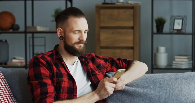 Potrait of the attractive hipster man with an earing sitting on the sofa in the living room and taping on the smartphone, then looking at the side. Indoor