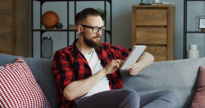 Caucasian male hipster with an earing and in the red motley shirt sitting on the sofa in the living room, taping on the tablet device, thinking and twisting his moustache. Inside