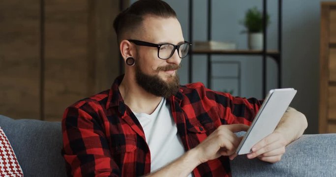 Portrait shot of the handsome hipster man with an earing and in the glasses sitting on the sofa in the living room, taping and typing on the tablet device. Indoors.
