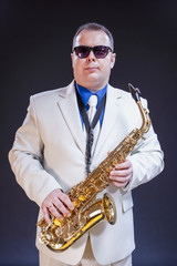 Confident Mature Male Saxophonist Posing with Instrument in White Suit and Sunglasses. Against Black Background.