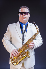 Naklejka premium Confident Mature Male Saxophonist Posing with Instrument in White Suit and Sunglasses. Against Black Background.