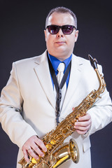 Fototapeta premium Music Themes. Portrait Of Confident Mature Male Saxophonist Posing with Instrument in White Suit and Sunglasses. Against Black Background.