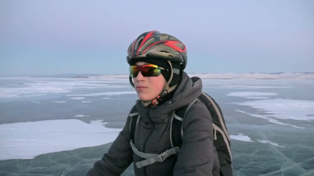 Man is stand a bicycle on ice. He looks at the beautiful sunset. The cyclist is dressed in a gray down jacket, backpack and helmet. Ice of the frozen Lake Baikal. The tires on the bicycle are covered