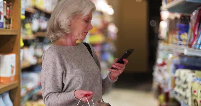 Senior woman looking at grocery aisle with cellphone in hand, Older woman using mobile phone app to shop for food, Retired person using diet app on smartphone in market, 4k 