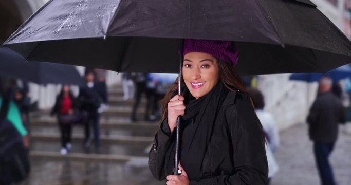 Smiling Latina woman taking shelter under umbrella, Portrait of young attractive woman outside on rainy day, 4k