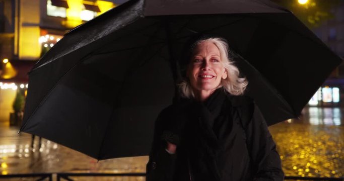 Senior woman in Paris dancing under umbrella on rainy night in the city, Beautiful mature Caucasian tourist waiting for ride in the rain, Retired female on vacation during winter, 4k