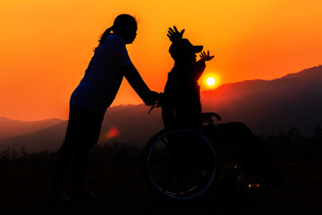 Silhouette of girl is helping man in Wheelchair strolling at sunset background