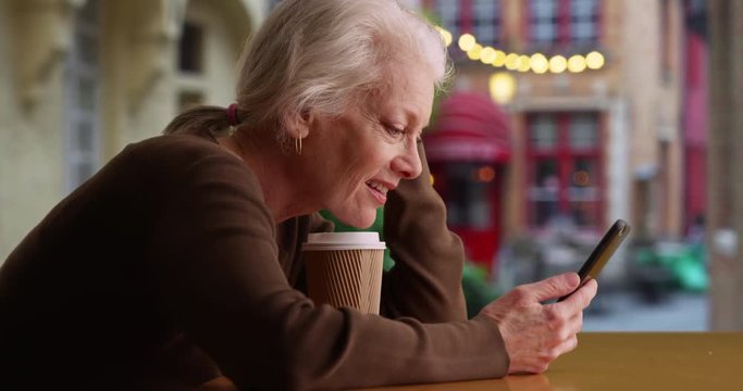 Smiling older Caucasian woman on cafe patio texting with friend on cellphone, Happy senior smiles as she sends and receives text messages at coffee shop using smart phone, 4k