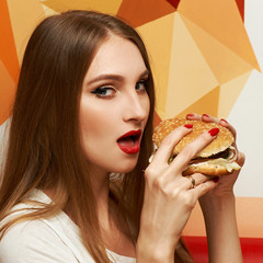 Portrait of gorgeous young woman with closed eyes and red lips holding tasty cheeseburger and biting it. Attractive female model eating burger with pleasure. Beautiful girl enjoying fast food meal.
