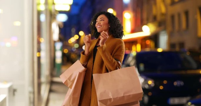 Stylish millennial black woman dancing and spinning with joy while shopping in the city at night, Excited young African American woman is happy with bags in her arms outside store, 4k