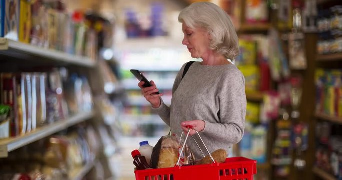 Mature woman looking at grocery aisle with smartphone in hand, Older woman using mobile phone app to shop for food, Retired person using diet app on smartphone in market, 4k 