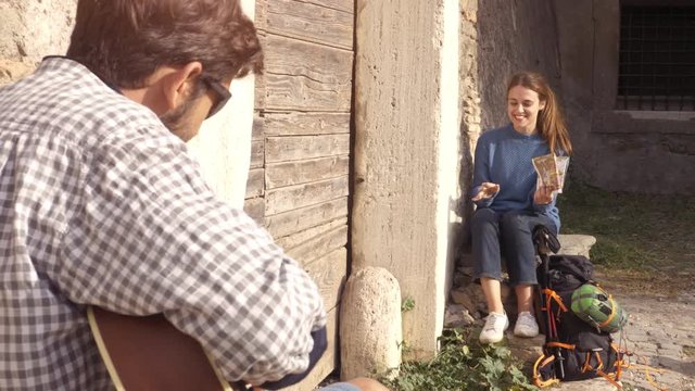 Happy young couple backpackers tourists playing guitar and singing sitting near wooden doorway of medieval building in parco degli acquedotti park ruins in rome at sunrise with map guide slow motion