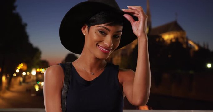 Stylish young black woman in Paris, France wearing elegant hat, Confident smiling African American female with her purse, posing for a portrait at night, 4k