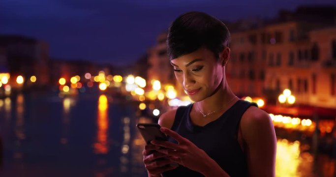 Pretty African American female traveling in Venice, Italy reads her phone messages smiling, Portrait of young black female with view of the Grand Canal using smartphone at night, 4k