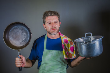 young attractive stressed and overwhelmed lazy man with apron holding kitchen pan and cook pot in...