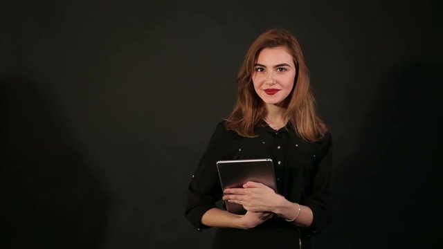 Young successful girl in a black business suit with a tablet in her hands on a black background. Portrait.