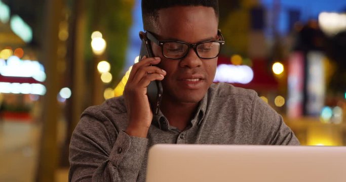 Happy millennial office worker chats on the phone on Champs-Elysees avenue, African American male sits with laptop talking on the phone at night, 4k