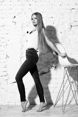 Young beautiful woman with long straight blonde hair in blue jeans and bright blouse standing against white brick wall and posing