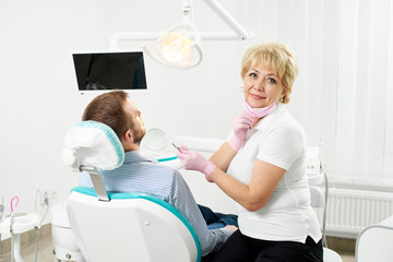 Obraz premium Modern clinic. Woman dentist looks into the camera and holding a tool, near her in the chair sits the patient