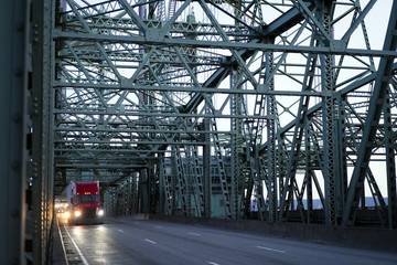 Red big rig semi truck with semi trailer running on interstate bridge in twilight