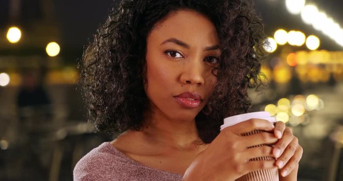 Close up of beautiful African American woman holding paper coffee cup outside at night, Black Millennial in the city on windy evening drinking out of to go cup and looking at camera, 4k