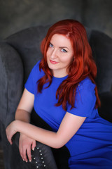 Fototapeta na wymiar Beautiful ginger-haired woman in blue dress sitting in gray armchair