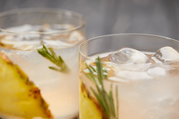 selective focus of two glasses of lemonade with pineapple pieces, ice cubes and rosemary on grey wooden tabletop