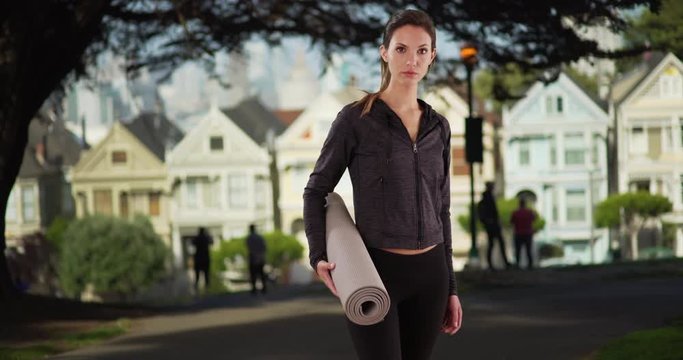 Woman with yoga mat walks to center screen and looks at camera near painted ladies homes, Portrait of pretty active yoga girl with mat walking at park in San Francisco, 4k