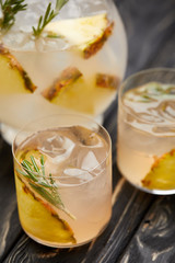 partial view of jug and two glasses of lemonade with pineapple pieces, ice cubes and rosemary on grey wooden tabletop