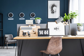 Brown leather chair standing by the wooden home office desk with lamp, poster and laptop in dark blue living room interior