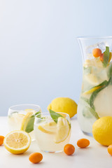 jug and two glasses of lemonade with mint leaves, ice cubes and lemon slices surrounded by kumquats and lemons on blue background